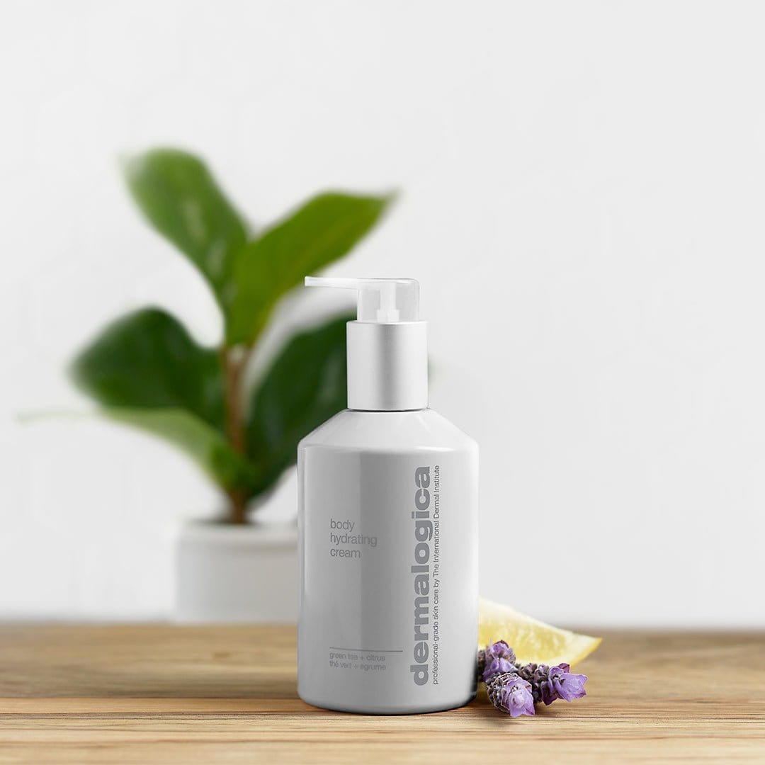 Eine weiße Pumpflasche mit Dermalogica Body Hydrating Cream steht auf einer weißen Oberfläche, im Hintergrund sind eine grüne Pflanze, lila Blumen und eine Zitronenscheibe zu sehen.