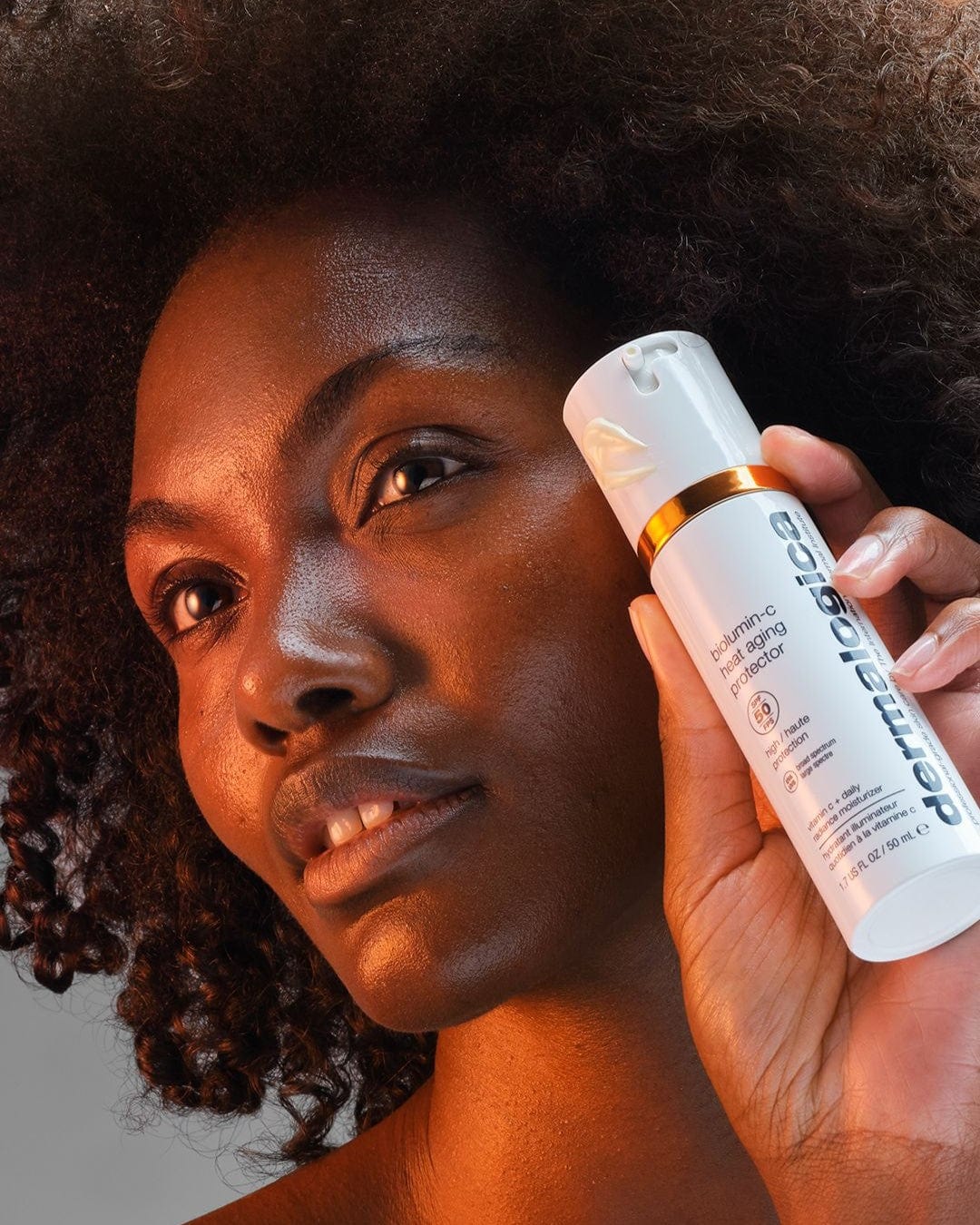 Eine Frau mit naturgelocktem Haar hält eine Flasche BioLumin-C Heat Aging Protector SPF50 + BioLumin C Serum dicht vor ihr Gesicht und zeigt sie stolz in die Kamera.