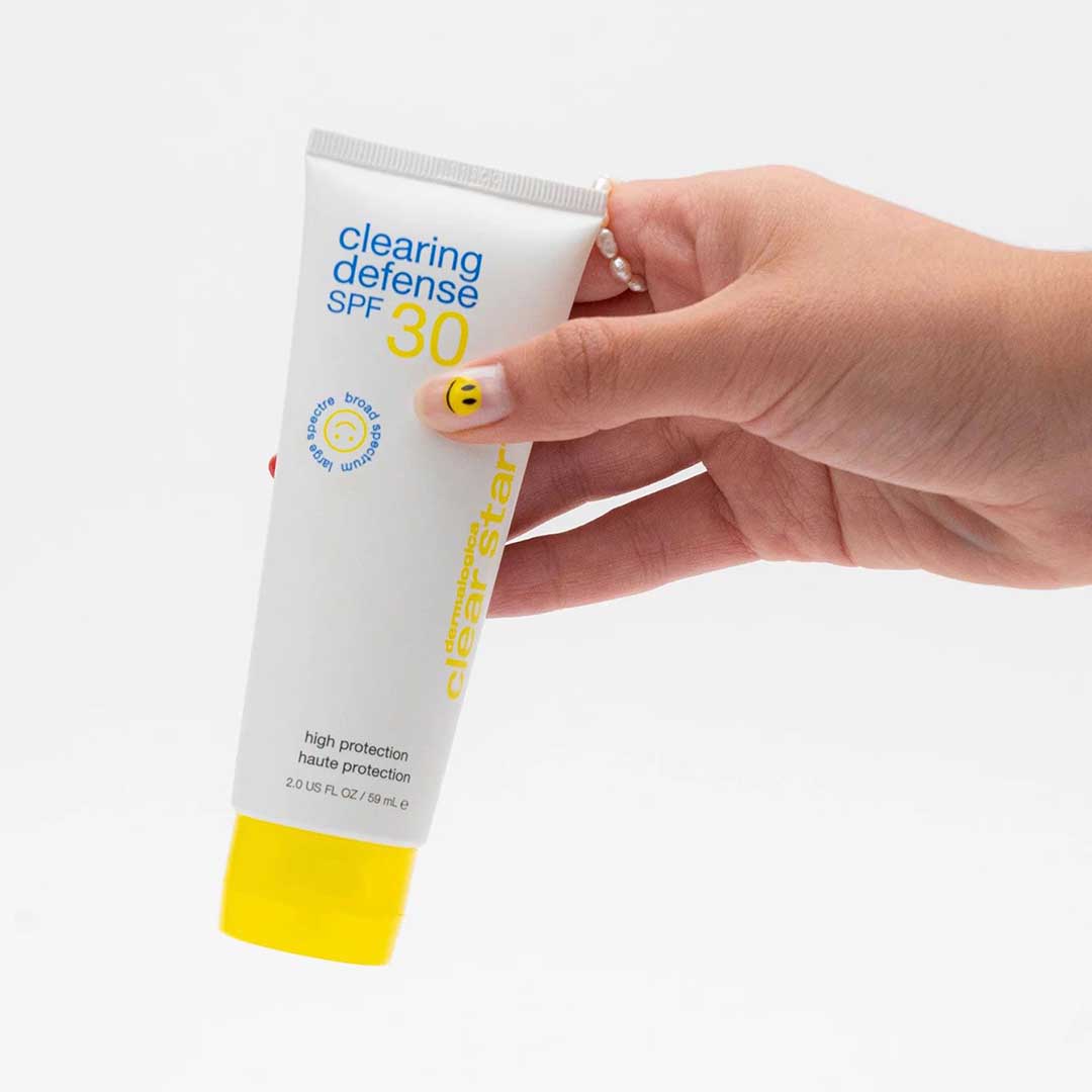 Eine Hand hält eine Tube Clearing Defense SPF30 mit gelbem Deckel vor weißem Hintergrund – eine ideale mattierende Feuchtigkeitscreme mit Breitband-UV-Schutz für fettige Haut.