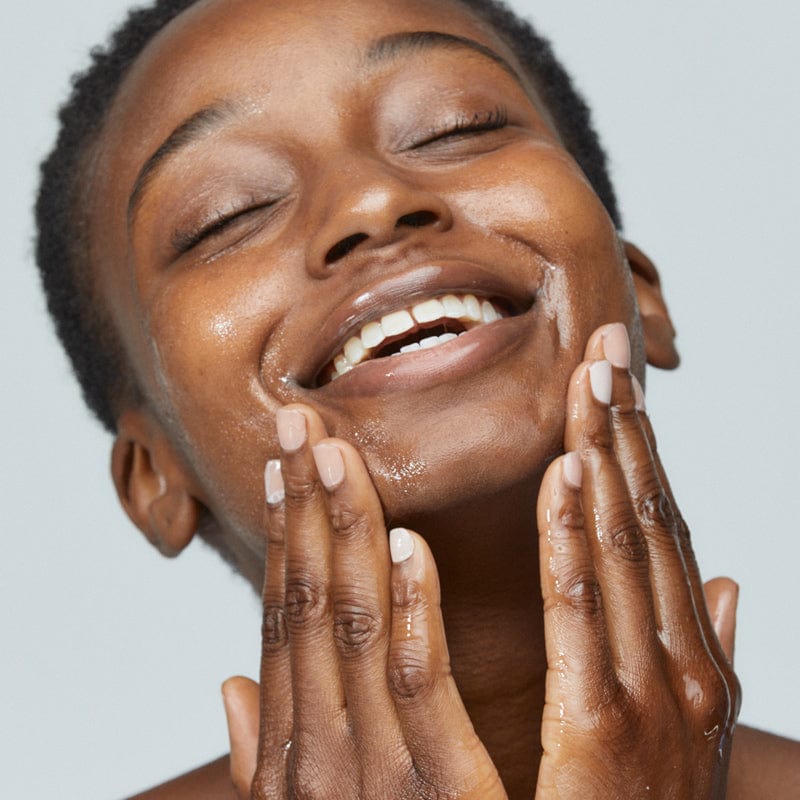 Eine Person mit feuchter Haut lächelt, während sie ihr Gesicht sanft mit beiden Händen berührt und eine Hautpflegeroutine mit PreCleanse als Make-up-Entferner vorschlägt.