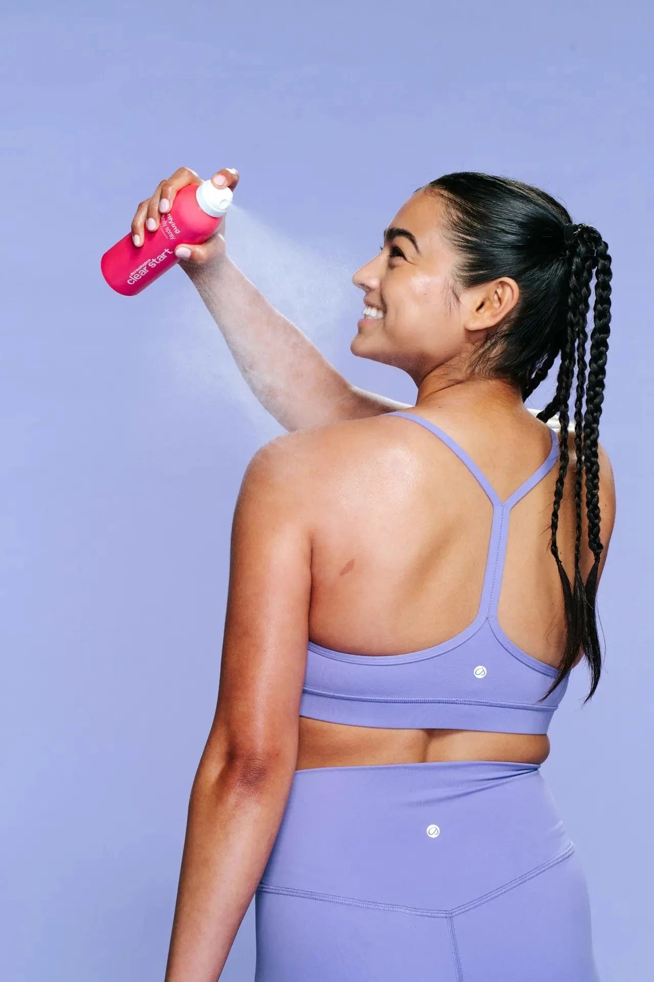 Eine Frau in einem lila Sportoutfit sprüht sich vor einem schlichten lila Hintergrund Clarifying Body Spray + Daily Microfoliant Sonnenschutz auf den oberen Rücken.