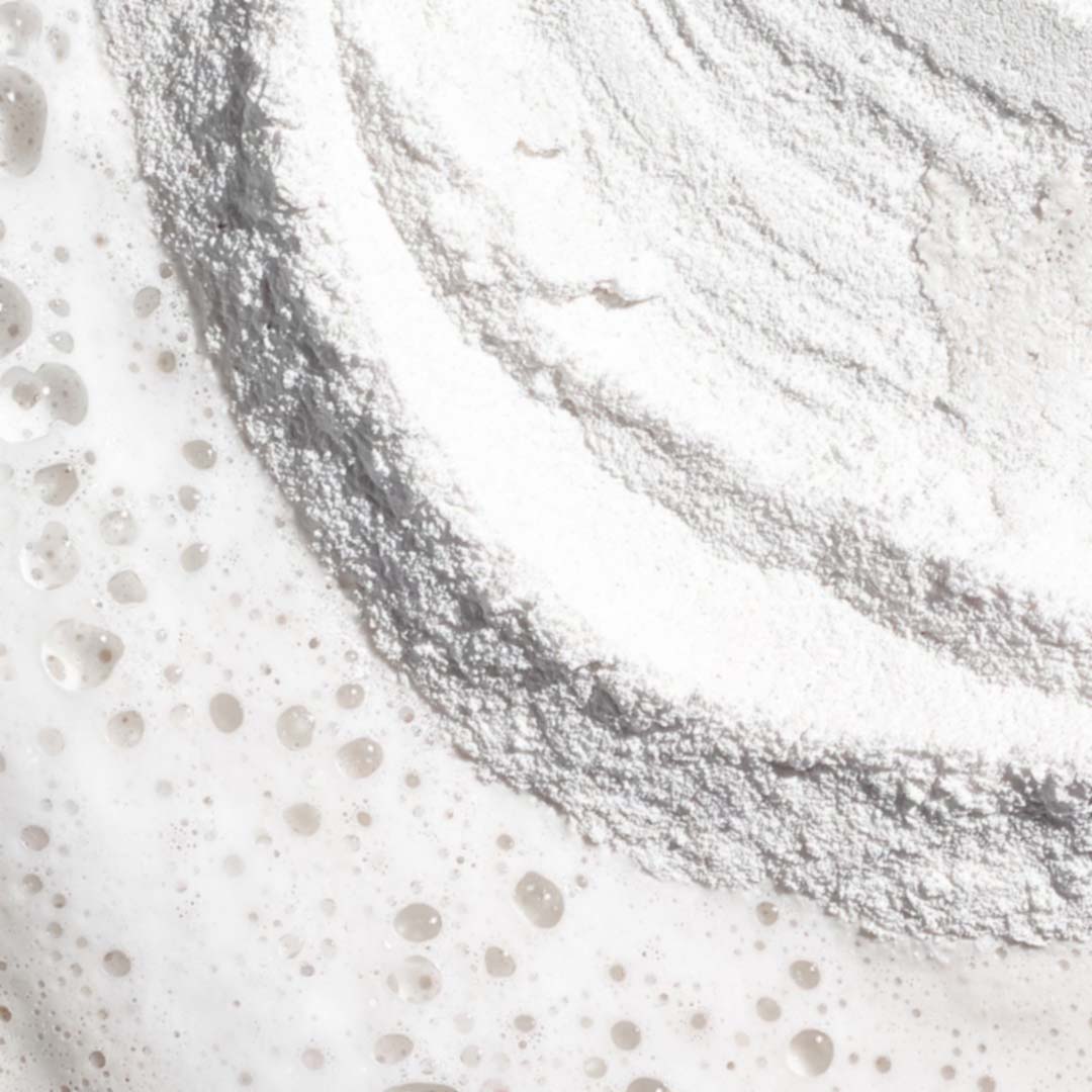 Nahaufnahme eines weißen Pulvers mit einem Ringmuster und verstreuten Flüssigkeitsblasen, das der feinen Textur von Daily Microfoliant ähnelt, einem Produkt zur Verfeinerung des Hautbildes.