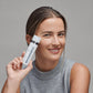 Eine Frau in einem grauen ärmellosen Oberteil lächelt und hält das PowerBright Dark Spot Serum + PowerBright Moisturizer SPF50, das sich ideal zur Behandlung von Pigmentflecken und Hyperpigmentierung eignet, vor einem schlichten grauen Hintergrund.