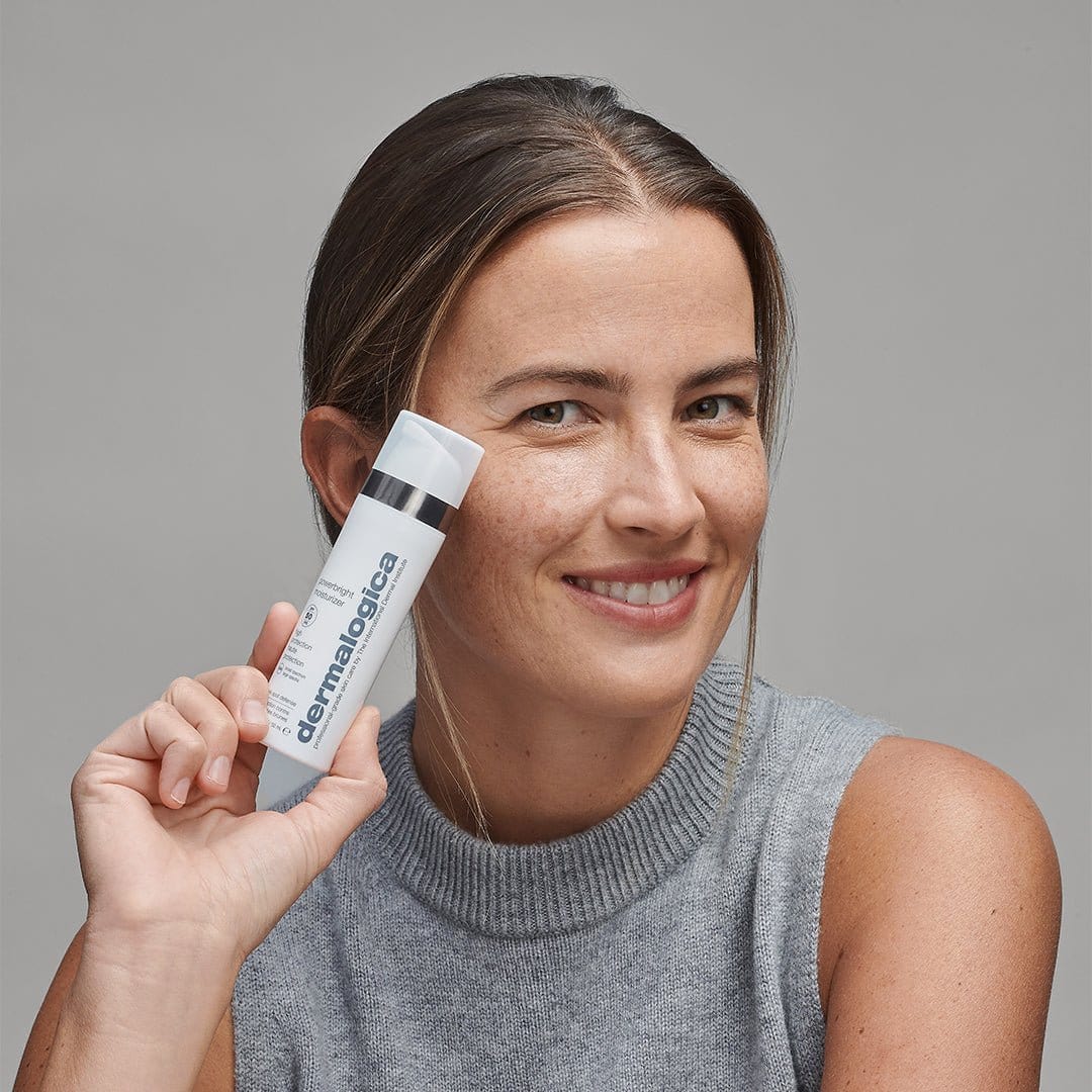Eine Frau in einem grauen ärmellosen Oberteil lächelt und hält das PowerBright Dark Spot Serum + PowerBright Moisturizer SPF50, das sich ideal zur Behandlung von Pigmentflecken und Hyperpigmentierung eignet, vor einem schlichten grauen Hintergrund.