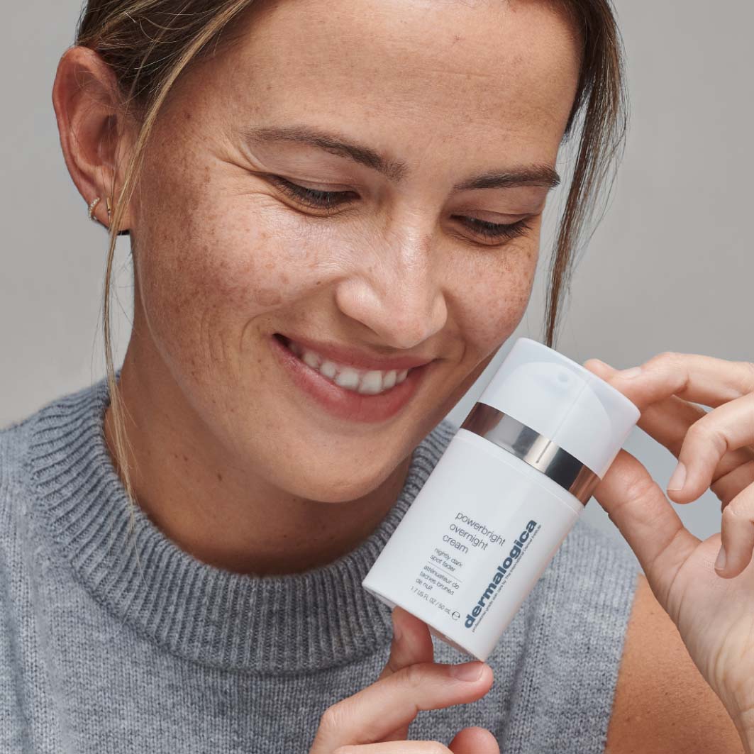 Lächelnde Frau hält ein Produkt von , die "PowerBright Overnight Cream", nah an ihr Gesicht.